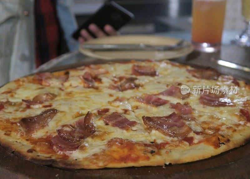 薄而脆的意式烟肉披萨配上金色融化的莫扎雷拉奶酪，在意大利餐厅露天用餐，在酒吧花园，晚上饥饿的用餐者拿着手机喝着啤酒/鸡尾酒，吃着美味的披萨